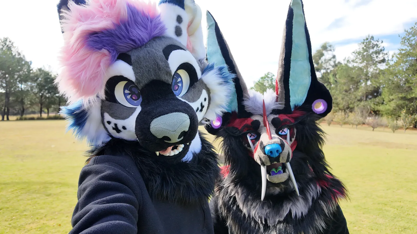 Selfie de Sheyko y Asmodeus en fursuit, en un parque: Sheyko con pelo rosa y morado, Asmodeus en negro y rojo con colmillos largos